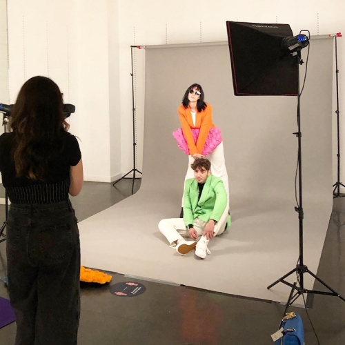 photographer taking an image of a person in a green jacket that's sitting on the floor and another person in an orange jacket standing over them